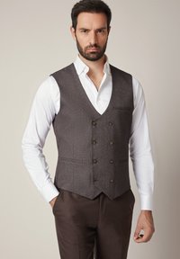 Brązowa kamizelka w jasnoprzecierane wzory houndstooth z dekoltem w kształcie litery V, podwójnym zapięciem z przodu, fakturowanym materiałem i dwoma kieszeniami; noszona na białej koszuli.