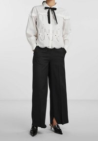 Camicia bianca di cotone con dettagli ricamati e motivi intagliati, con un nastrino nero. Abbinata a pantaloni neri a gamba larga.
