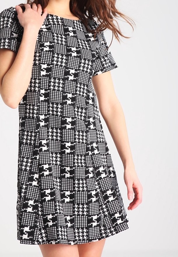 Femme portant une robe à manches courtes à carreaux noirs et blancs avec des motifs géométriques variés sur un fond uni.