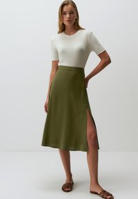 Jupe midi vert olive avec une fente sur le côté, fabriquée en tissu léger. Associée à un haut ajusté en maille blanche et des sandales à glisser brunes.