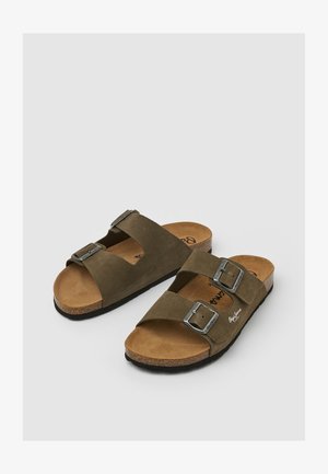 Para brązowych zamszowych sandałów z podwójnymi, regulowanymi metalowymi klamrami i korkowymi podeszwami na jasnoszarym tle.