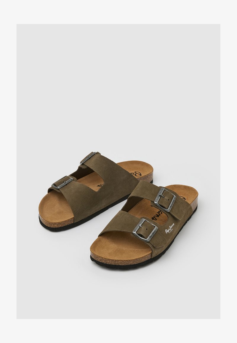 Coppia di sandali in suede marrone con doppia fibbia regolabile in metallo e plantari in sughero su uno sfondo grigio chiaro.