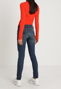 Jeans de denim oscuros de corte ajustado con bolsillos traseros, combinados con una camiseta de manga larga brillante de color naranja acanalada y zapatillas blancas.