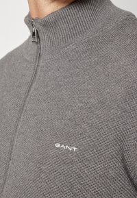 Gros plan d'une personne portant un pull zippé gris en maille avec un col montant et le logo blanc GANT sur la poitrine.
