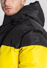 Pufjakke med sort overdel og gul underdel, lavet af shiny nylon. Indeholder et logo-patch på venstre skulder.