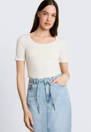 Jonge vrouw met lang bruin haar die een wit geribd T-shirt met korte mouwen draagt en een lichtblauwe high-waisted spijkerrok met een gestrikte riem.