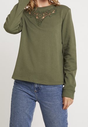 Kobieta w oliwkowozielonym, długim rękawie z kwiatową koronką przy dekolcie i niebieskich dżinsach, stojąca na jednolitym tle.