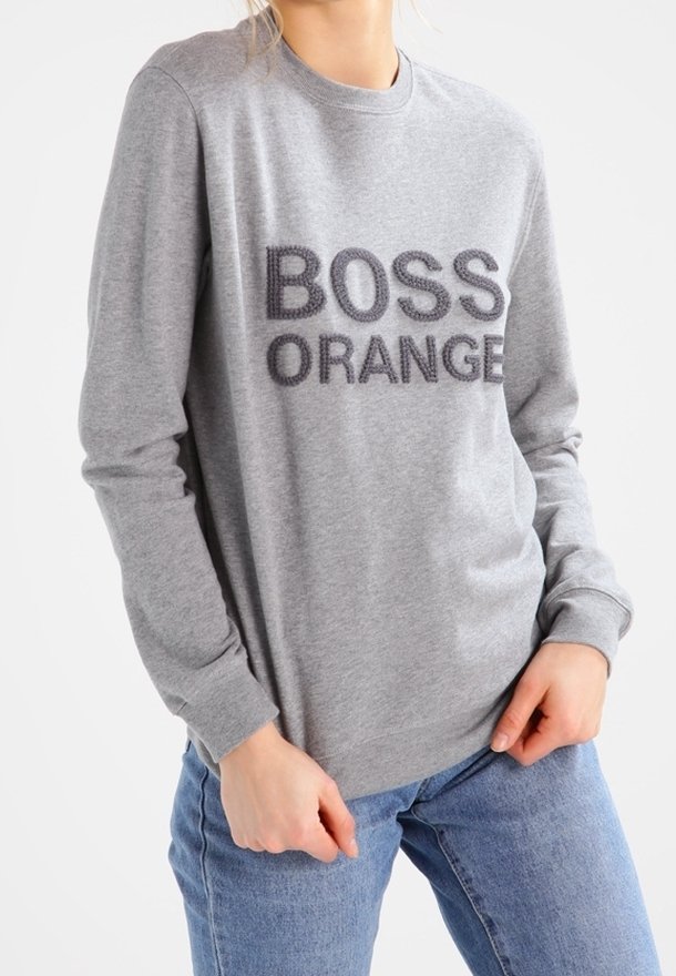 Personne portant un sweat-shirt gris avec le texte "BOSS ORANGE" et un jean bleu, posant les mains près de la taille.