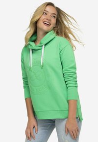 Sudadera verde con un logotipo circular texturizado en el frente, cuerdas blancas y un zipper lateral. Hecha de material suave de sudadera.