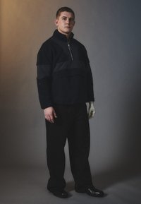 Zwarte fleece pullover met een halve rits, voorzien van een donkere stoffen overlay, losse pasvorm en elastische manchetten, gecombineerd met brede zwarte broeken en schoenen.