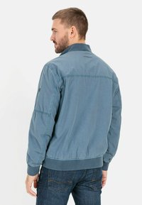 Lichtblauwe jas met een ontspannen pasvorm, voorzien van een kraag, ribgebreide manchetten en stikseldetails. Draagt over donkere jeans.
