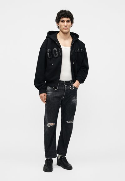 Νέος άντρας στέκεται φορώντας μαύρο ζακέτα με φερμουάρ και κείμενο, λευκό φανελάκι, φθαρμένα μαύρα τζιν με μεταλλικούς κρίκους και μαύρα αθλητικά παπούτσια σε λευκό φόντο.