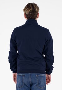 Giacca a vento blu navy con colletto alto, polsini e orlo a coste. Il materiale sembra liscio con una leggera lucentezza. Indossata con jeans blu.