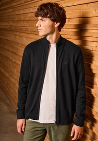 Černý svetr na zip vyrobený z měkké látky, s žebrovaným límcem a manžetami. Nositý přes bílou košili, v kombinaci s olivově zelenými kalhotami.