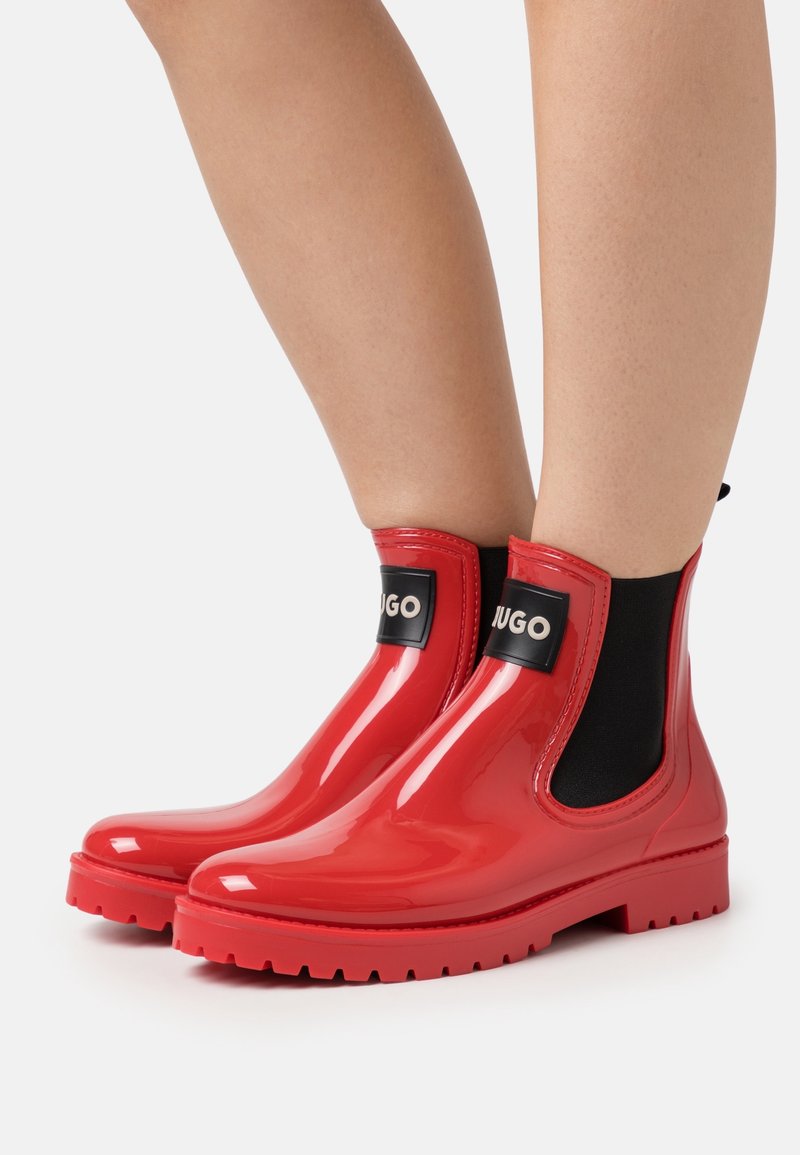 Bottes Chelsea en caoutchouc rouge avec une finition brillante, panneaux élastiques sur les côtés et accents texturés noirs arborant un logo de marque. Semelle plate à crampons.
