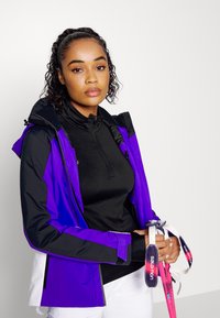 Damen-Skijacke in Schwarz und Lila mit durchgehendem Reißverschluss und verstellbarem Kapuzen. Sie hält pinkfarbene Skistöcke mit bedruckten Schlaufen.