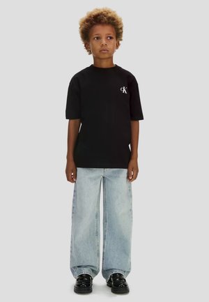 Bambino con capelli ricci indossa una maglietta nera CK, jeans larghi azzurri e scarpe nere su uno sfondo semplice.