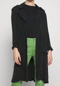 Svart lång kappa med en avslappnad passform, öppen front och stora sidofickor, bärs över en svart skjorta och gröna byxor. Slät tygkvalitet.