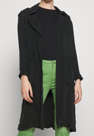Abrigo negro largo con un corte relajado, abierto por delante y grandes bolsillos laterales, usado sobre una camisa negra y pantalones verdes. Textura de tela suave.
