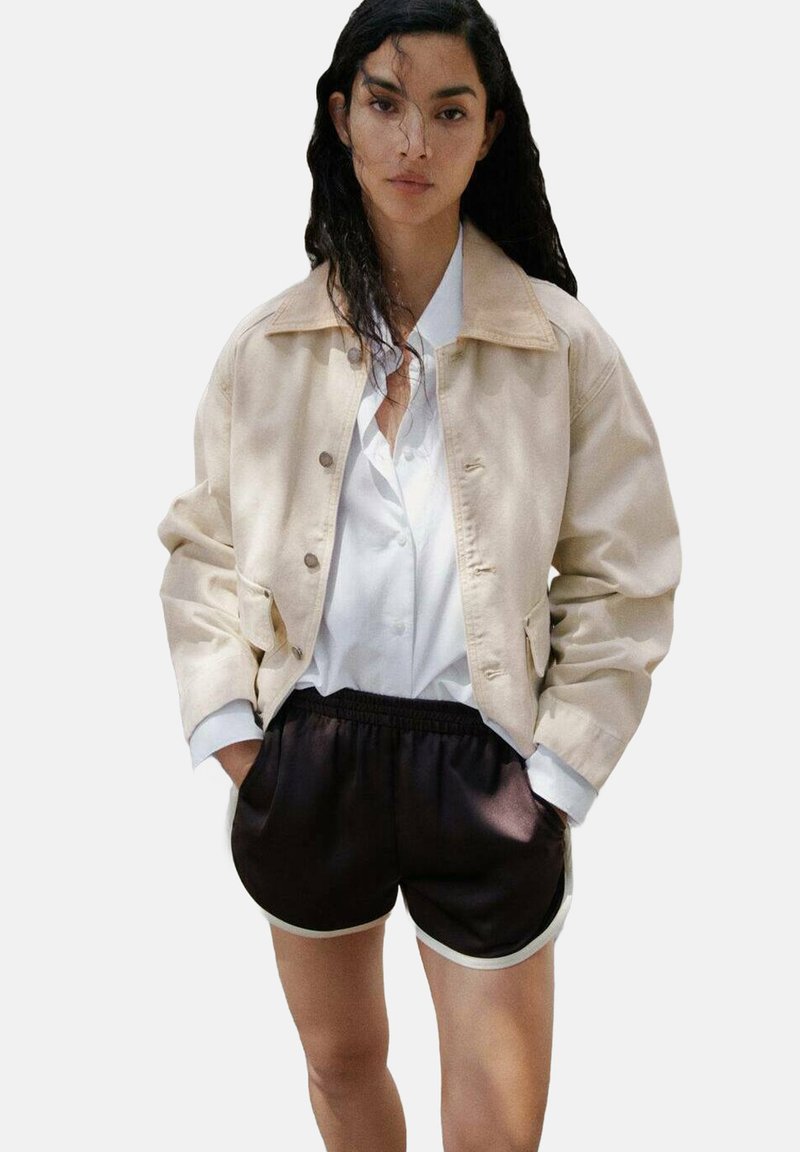Veste en denim beige avec un col, portée sur une chemise blanche à boutons. Shorts en satin noir avec des bordures blanches. Coupe décontractée et design casual.