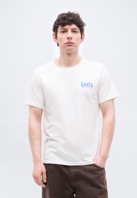 Νεαρός με σγουρά μαλλιά, φορώντας λευκό μπλουζάκι Levi’s και σκούρο παντελόνι, στέκεται με το ένα χέρι στην τσέπη μπροστά σε απλό φόντο.