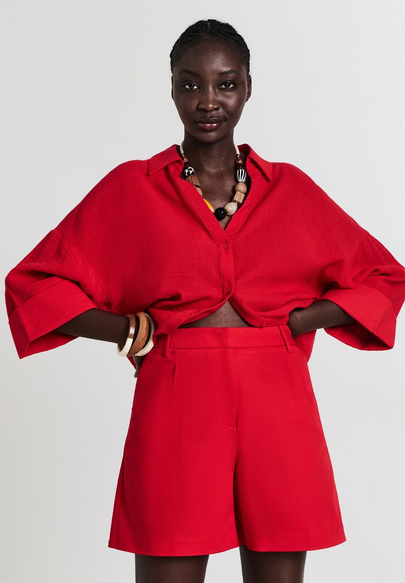 Donna che indossa una camicia rossa larga con bottoni e pantaloni corti rossi a vita alta, con una collana di perline multicolori e braccialetti di legno.