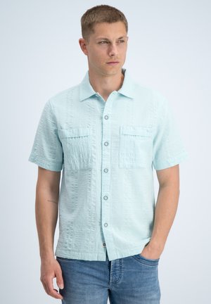Junger Mann trägt hellblaues strukturiertes Kurzarmhemd mit Fronttaschen und blaue Jeans, schaut zur Seite, eine Hand in der Tasche.