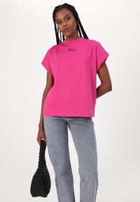 Pink short-sleeve t-shirt with a high neckline and embroidered logo. Paired with light gray jeans and a black woven handbag.
