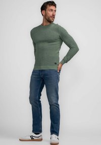 Maglione verde a coste con maniche lunghe, abbinato a jeans blu e sneakers bianche. Il maglione presenta un tessuto testurizzato e un logo discreto.