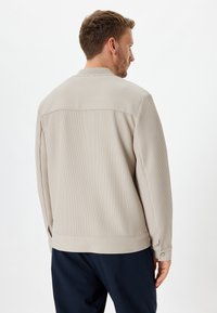Beige stribet jakke med en afslappet pasform, der har en rund halsudskæring, lange ærmer og manchetter med knapper. Tekstureret stof med synlige søm detaljer.