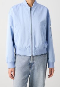 Jasnoniebieska kurtka bomber z zamkiem błyskawicznym, ściągaczami na mankietach i u dołu oraz dwoma przednimi kieszeniami; posiada małą kieszeń na zamek na rękawie.