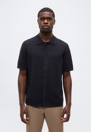 Jeune homme debout face à l’objectif, portant une chemise noire à manches courtes avec col et un pantalon beige sur un fond clair uni.