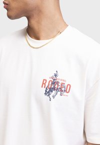 Biała bawełniana koszulka z graficznym nadrukiem rodeo jeźdźca i byka w niebieskim i czerwonym kolorze, z napisem "REDEFINED REBEL" poniżej.