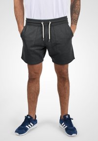 Shorts de sport gris avec une taille élastique, des poches avant et un cordon de serrage ; portés avec des chaussures de sport bleues comportant des accents blancs.