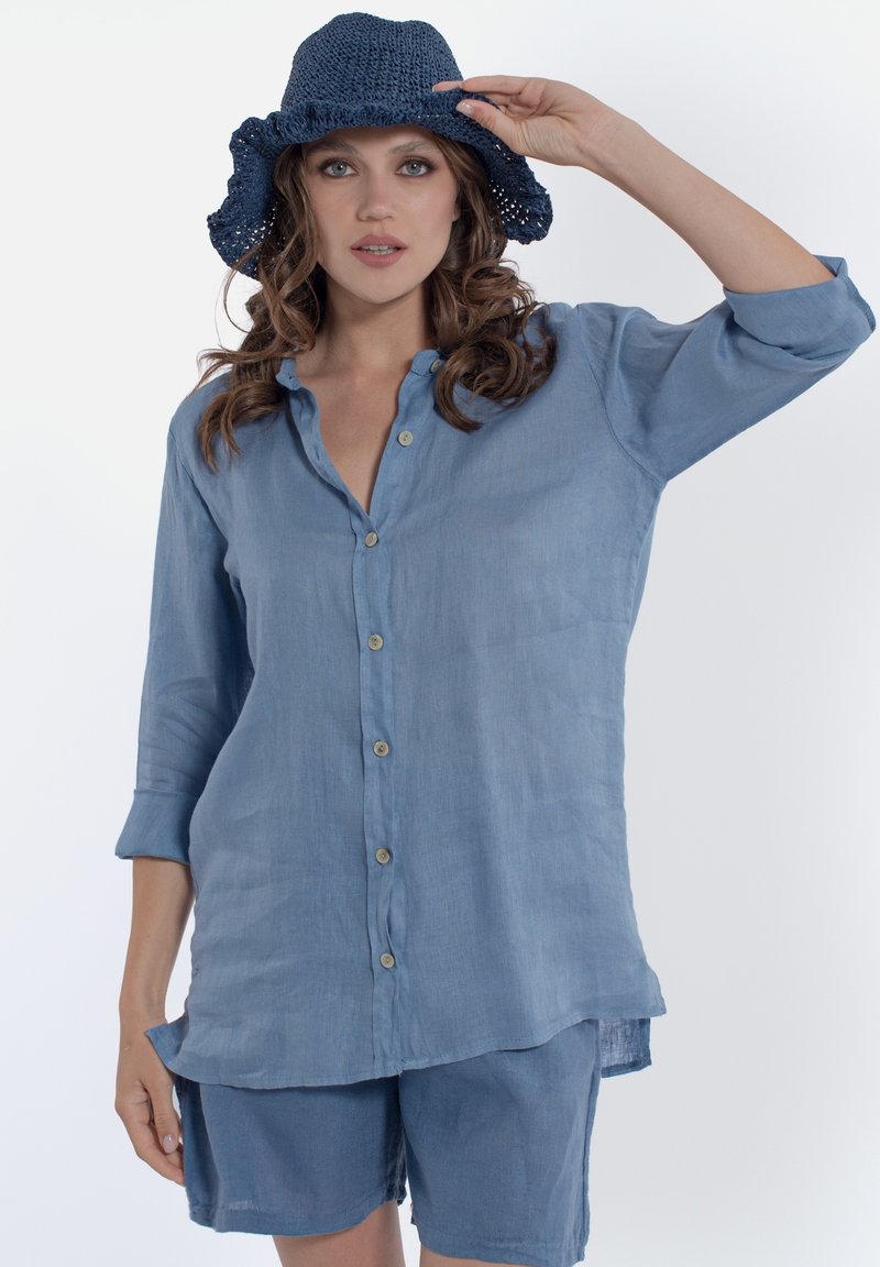 Camicia di lino azzurro chiaro con design a bottoni, maniche arrotolabili e pantaloni corti abbinati. Il modello indossa un cappello a secchiello azzurro all'uncinetto.