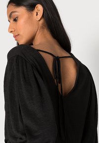 Top preto texturizado com um decote em V profundo nas costas, com cintas amarradas. As mangas bufantes oferecem volume, destacando uma silhueta descontraída, mas estruturada.