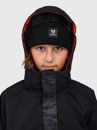Niño con una chaqueta de invierno negra con capucha y un gorro de lana negro con el logo de Brunotti, sobre un fondo gris.