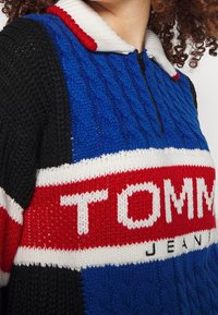 Pull en maille avec texture torsadée en bleu, manches noires et col blanc. Large bande rouge et blanche avec le logo "TOMMY JEANS". Détail zippé.