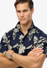 Camisa de manga corta de color navy con suaves patrones florales, cuello abotonado y tela suave. Reloj dorado con correa de cuero marrón claro en la muñeca.