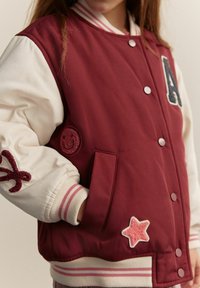 Burgundia värvi varsity jakk valgete varrukatega, millel on täht "A", naeratav õmblus ning roosa tähe aksent. Ribidega mansetid triipudega.