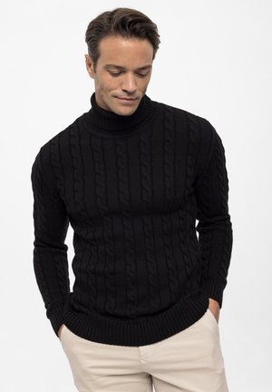 Homme aux cheveux courts et foncés portant un pull à col roulé noir en maille torsadée et un pantalon beige, regardant vers le bas avec les mains dans les poches.