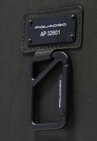 Zwarte leren label met een zilveren "Piquadro" logo en de tekst "AP 32601", met een zwarte metalen karabijnhaak eraan bevestigd. Getextureerde achtergrond.