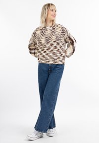 Kreminės ir rudos spalvos megztinis, laisvo kirpimo, su plačiomis rankovėmis ir tekstūruotu raštu, derinamas su mėlynomis plačiomis kelnėmis ir baltomis sportbačiais.