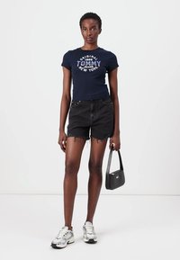 Tamsiai mėlyna prigludusi marškinėliai su "Original Tommy Jeans New York" grafika, derinami su aukšto juosmens juodais džinsiniais šortais ir sidabriniais sportbačiais.