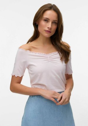 Donna con capelli castano chiaro che indossa una maglia rosa pallido con spalle scoperte e una gonna di jeans azzurro chiaro, in piedi davanti a uno sfondo bianco uniforme.