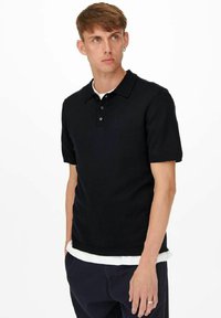 Zwarte polo met korte mouwen en kraag, vierknopenboord, ribgebreide manchetten en een getailleerd ontwerp, gedragen over een wit hemd.
