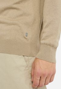 Beige kötött pulóver bordás végződéssel és mandzsettával. Az oldalán egy kis fém logó található, világos bézs nadrággal párosítva.