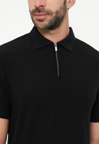 Homme portant un polo noir en maille à manches courtes avec une fermeture éclair argentée au col, montrant un motif losange texturé sur le tissu.