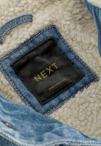 Etiqueta de tela negra con "NEXT" y "Hecho en Bangladés" cosida en una prenda de mezclilla azul y forro de lana beige.