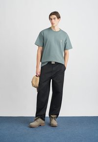 Grøn bomulds t-shirt med et lille logo, sorte vide bukser, beige kasket og grå fodtøj med en struktureret sål, sat mod en lys baggrund.